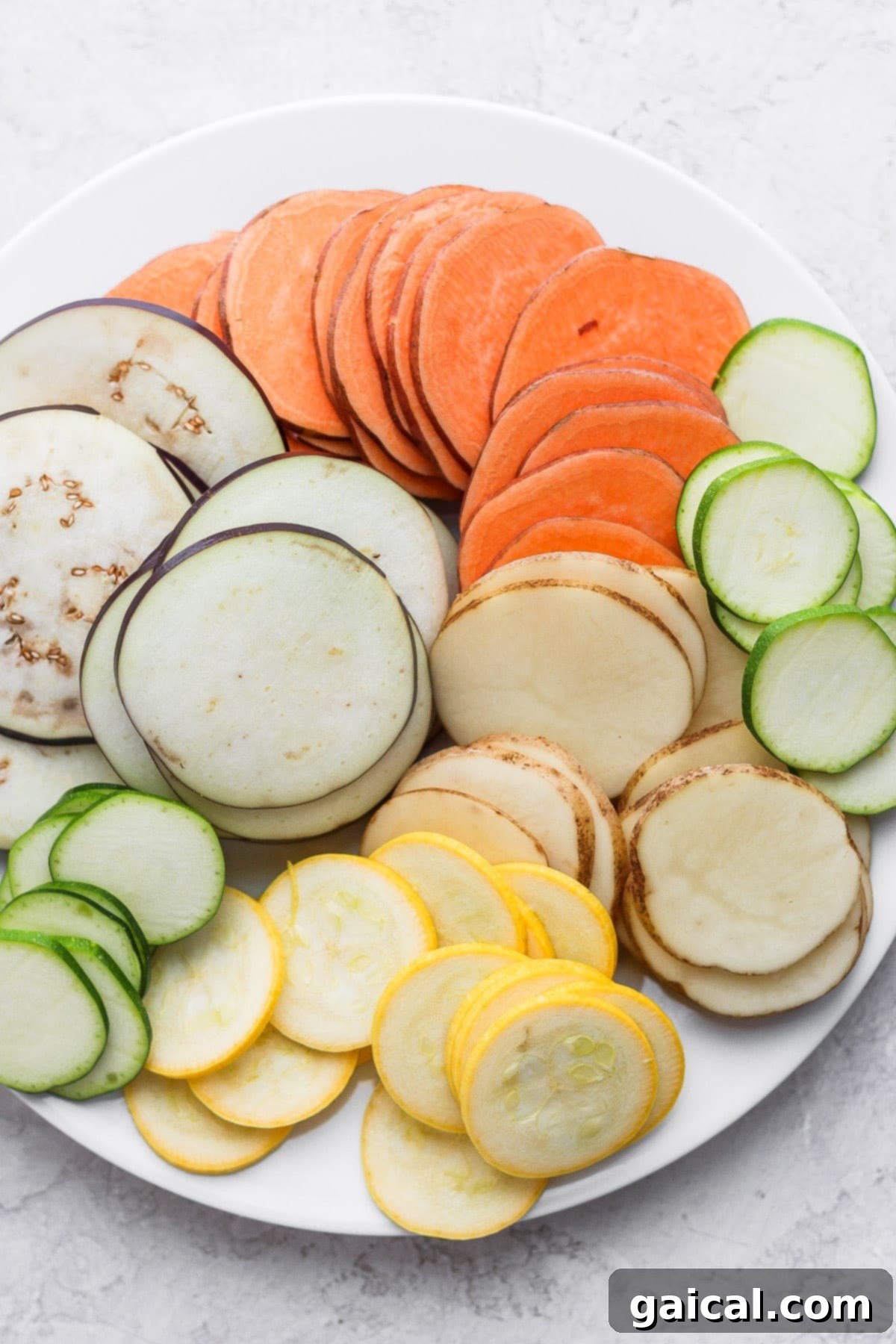 Freshly sliced root vegetables, including eggplant, zucchini, and potatoes, arranged on a plate.