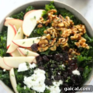 The Ultimate Thanksgiving Salad 5 A large bowl of Thanksgiving salad, featuring massaged kale, apple slices, candied walnuts, and goat cheese, with dressing drizzled over.
