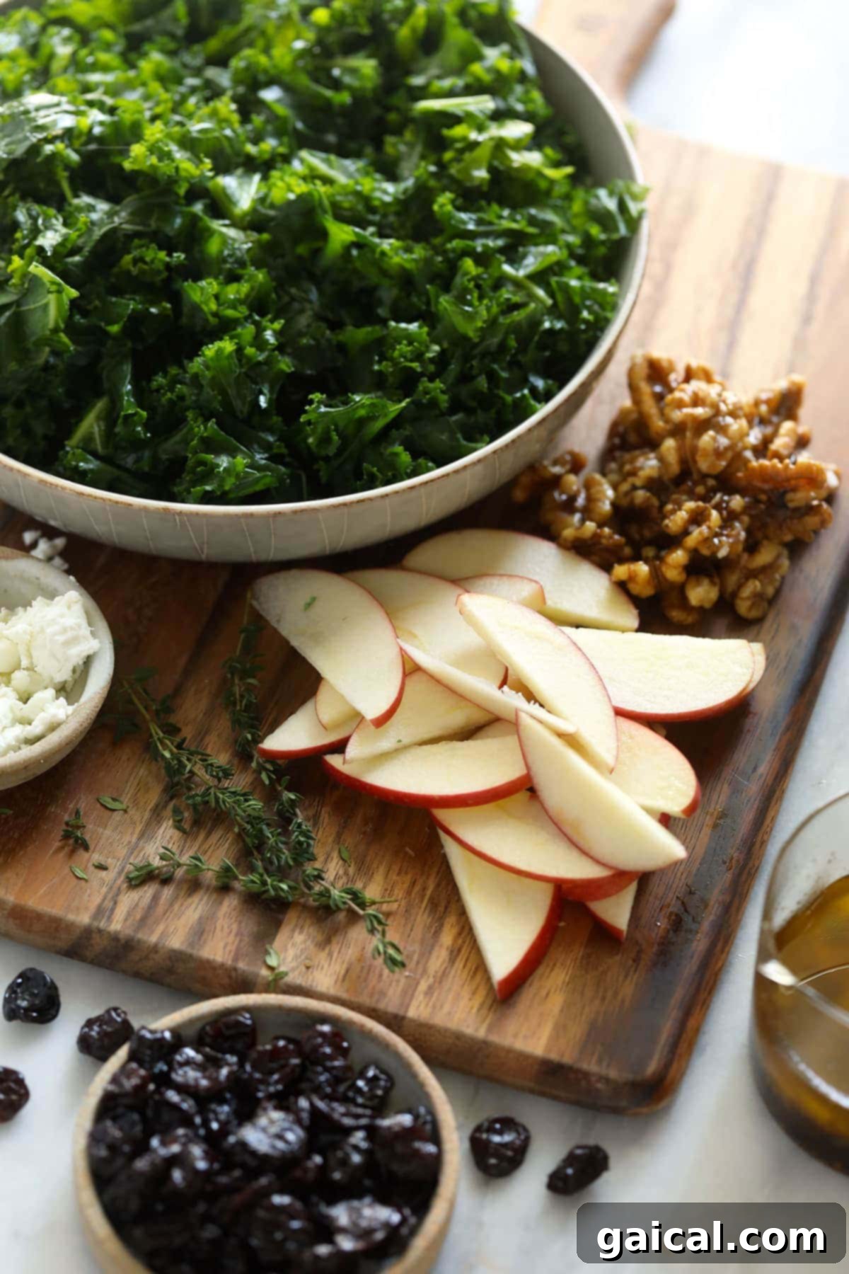 Looking for a last-minute Thanksgiving salad idea? Try our delicious massaged kale, apple, and walnut salad made with goat cheese, dried cherries, and our favorite vinaigrette! Various ingredients for a Thanksgiving salad laid out on a wooden cutting board, including kale, apples, goat cheese, candied walnuts, and dried cherries.