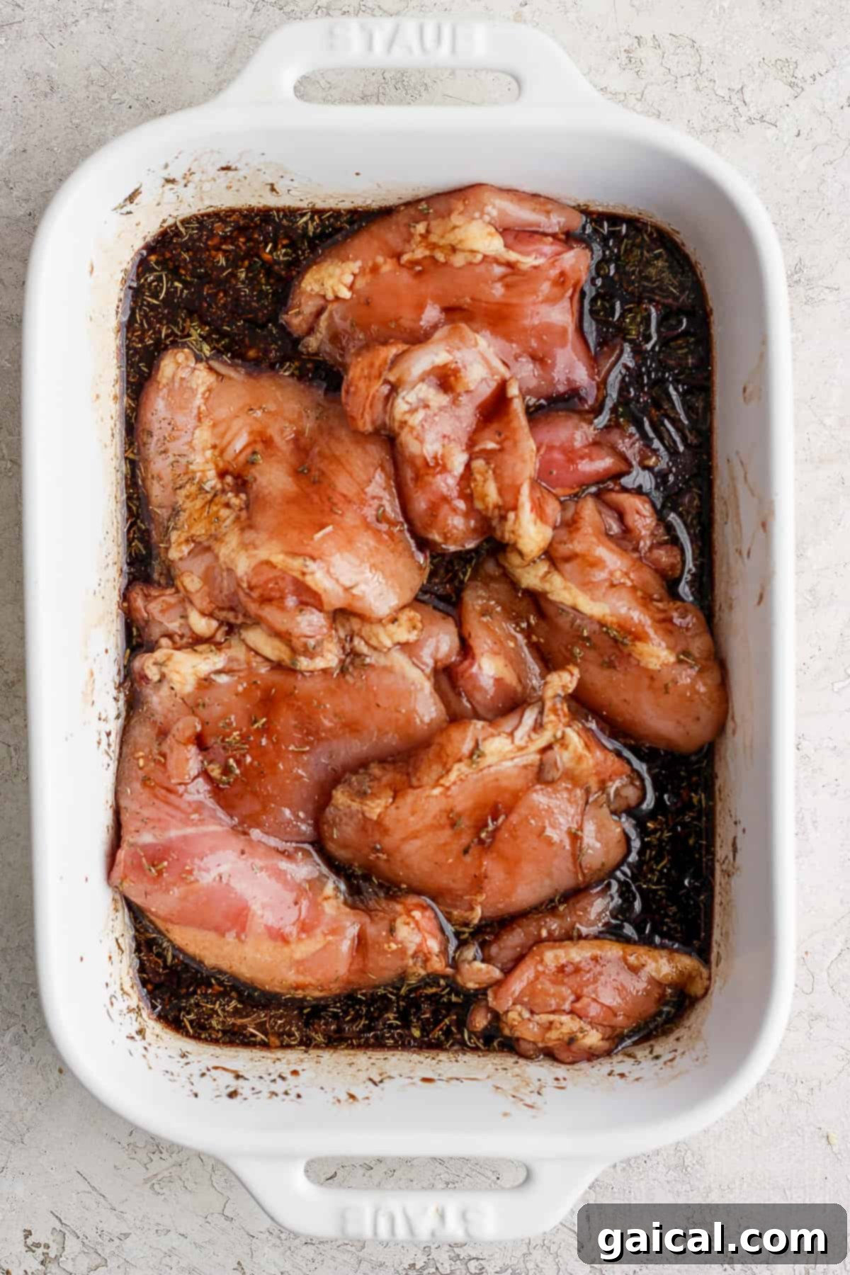 Raw chicken breasts completely covered in a balsamic marinade in a white baking dish, sealed with plastic wrap for marinating.