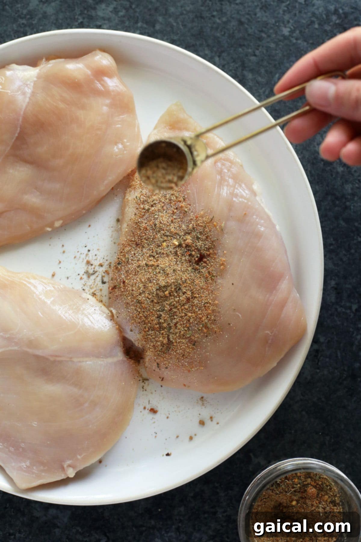 Universal Poultry Flavor Blend 2 A chef's hand generously sprinkling all-purpose chicken seasoning over a raw chicken breast, ensuring an even and thorough coating on a wooden cutting board.