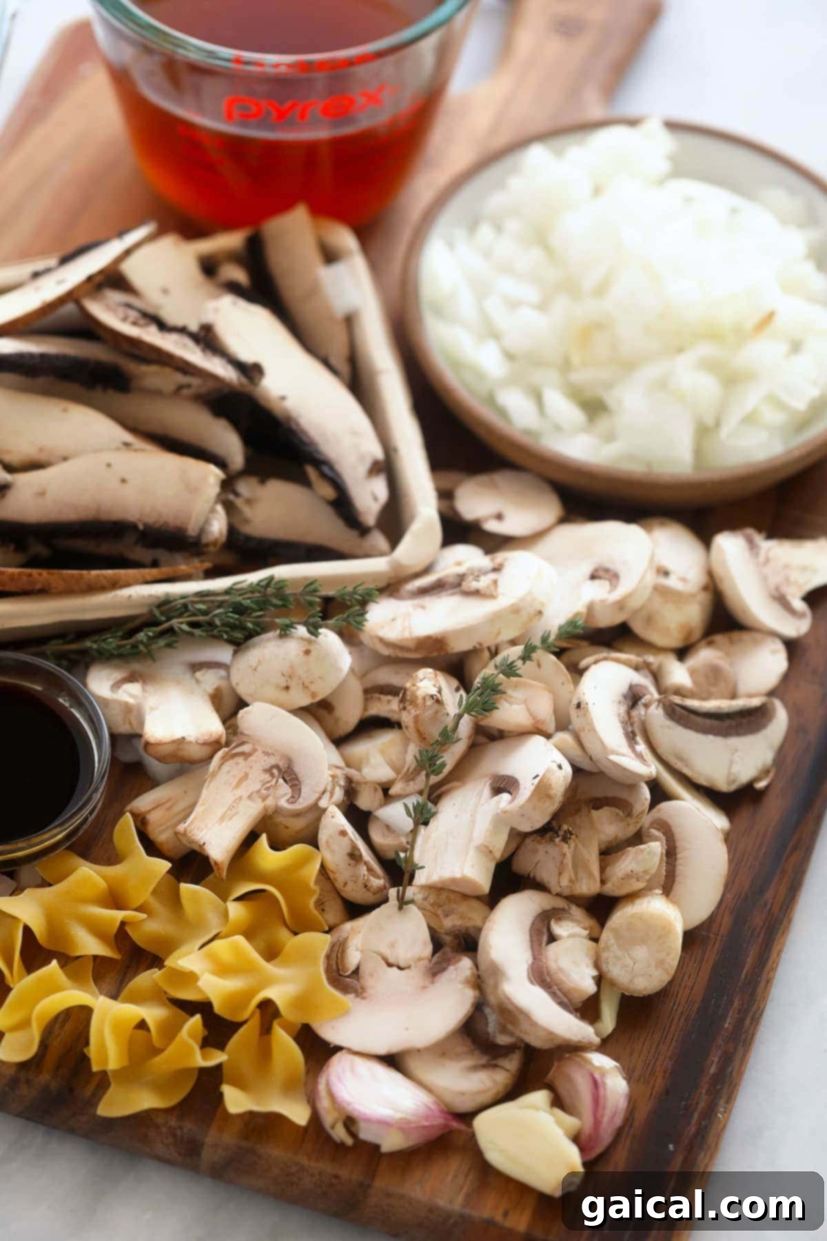 Creamy Mushroom Stroganoff 3 An assortment of fresh ingredients laid out on a wooden cutting board for a healthy mushroom stroganoff, including various types of mushrooms, a diced white onion, garlic cloves, a measuring cup of broth, and fresh thyme sprigs.