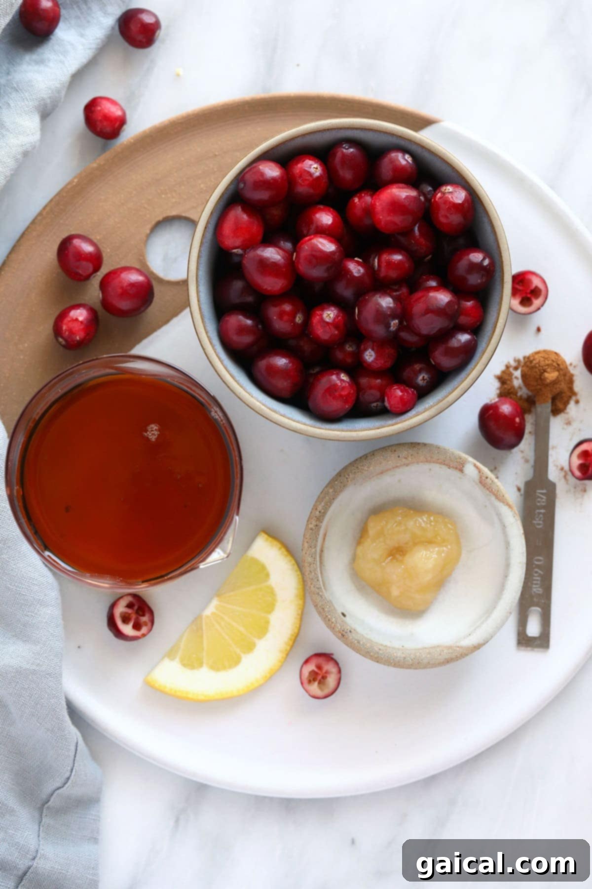 Arrangement of fresh ingredients for lemon ginger cranberry sauce: cranberries, maple syrup, grated ginger, lemon, and cinnamon stick on a white plate.