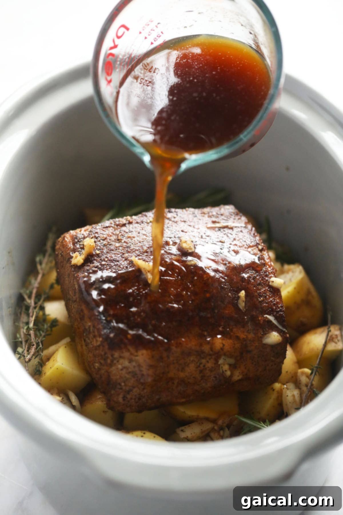 This is the most tender Slow Cooker Pork Loin roast recipe ever! It's served with potatoes and a savory gravy for the perfect meal. Enjoy! pork loin roast on a bed of diced potatoes and aromatic herbs being carefully placed into a slow cooker.