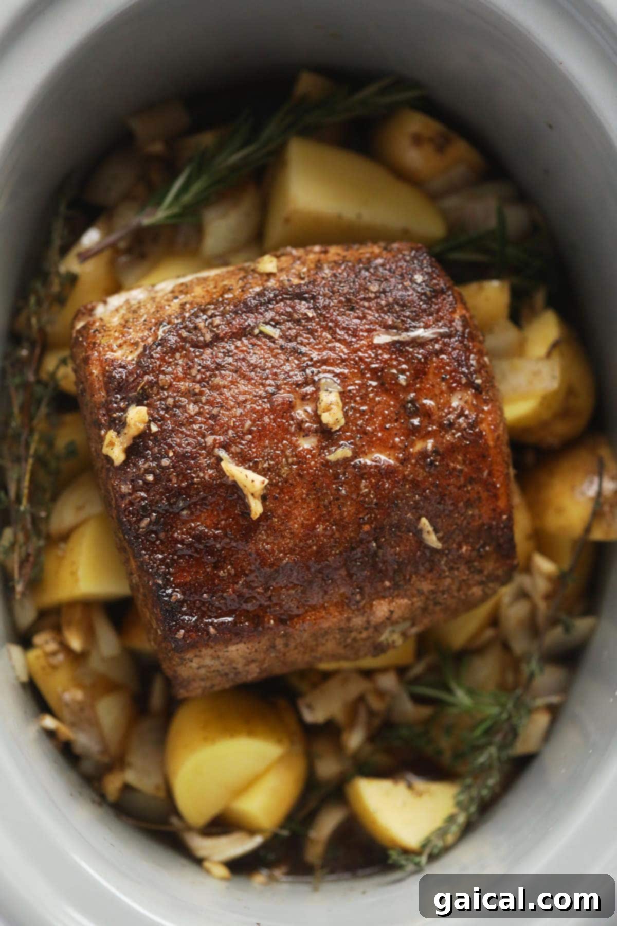 This is the most tender Slow Cooker Pork Loin roast recipe ever! It's served with potatoes and a savory gravy for the perfect meal. Enjoy! slow cooker pork loin roast on a bed of potatoes and fresh herbs in the slow cooker, ready to serve.