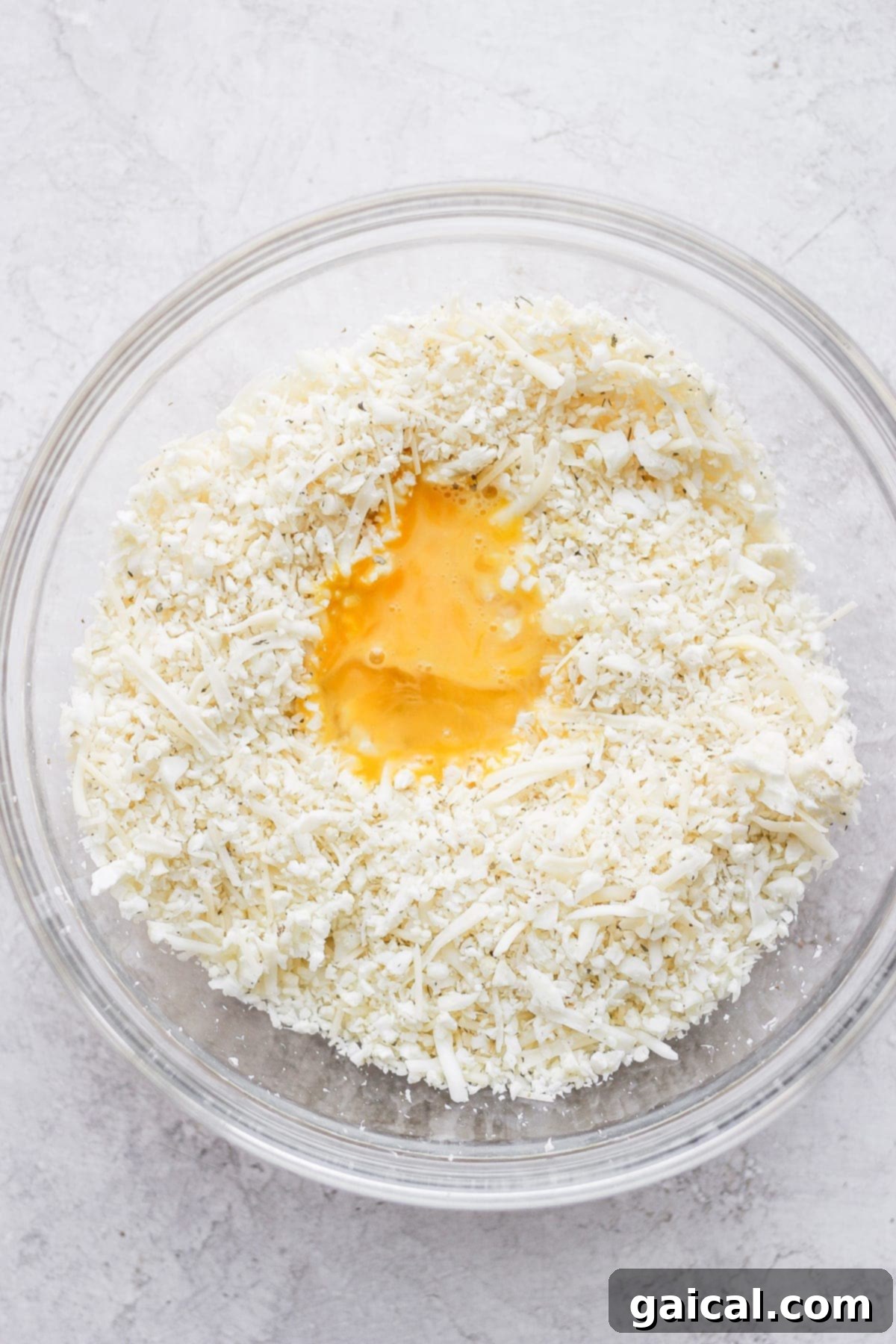 Bowl containing cauliflower rice, shredded cheeses, and a whisked egg, all mixed together to form the pizza crust dough.