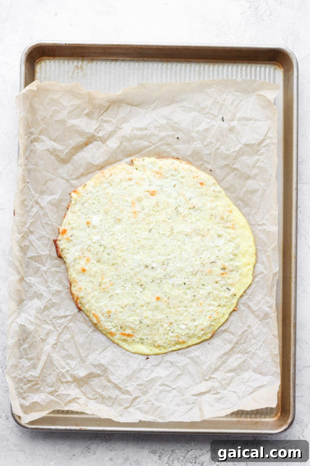 Partially baked cauliflower pizza crust, golden brown at the edges, resting on parchment paper after its initial bake.