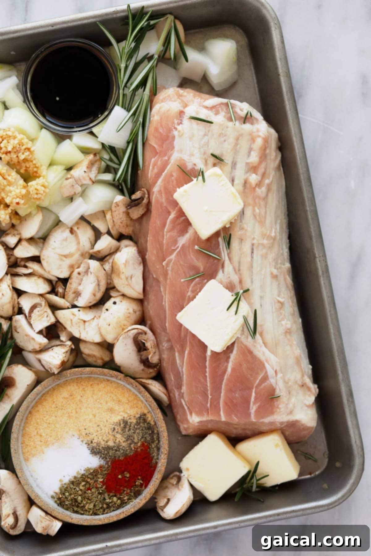 Raw pork tenderloin seasoned with spices, alongside sliced mushrooms and butter on a sheet pan.