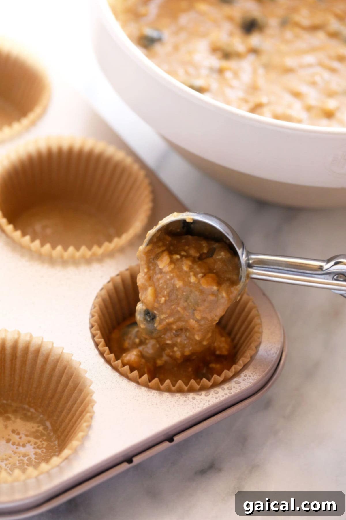 Blueberry oatmeal muffin batter being carefully spooned into paper-lined muffin cups in a baking tin.