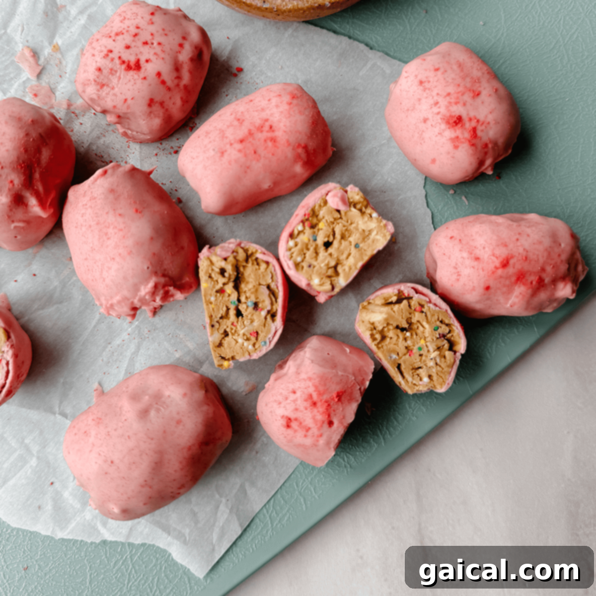 Cake Batter Protein Bites 5 Pink-coated truffles on parchment paper, some sliced open to reveal a cake-batter-like filling with colorful sprinkles, resembling Easter eggs.