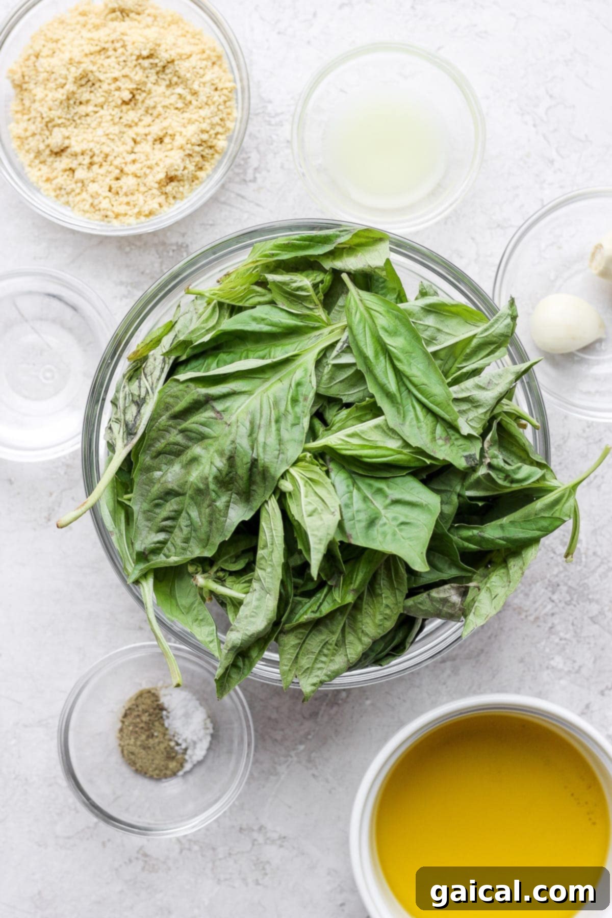 Array of fresh vegan pesto ingredients laid out: fresh basil, garlic cloves, whole lemon, a bowl of vegan parmesan cheese, and a bottle of olive oil.