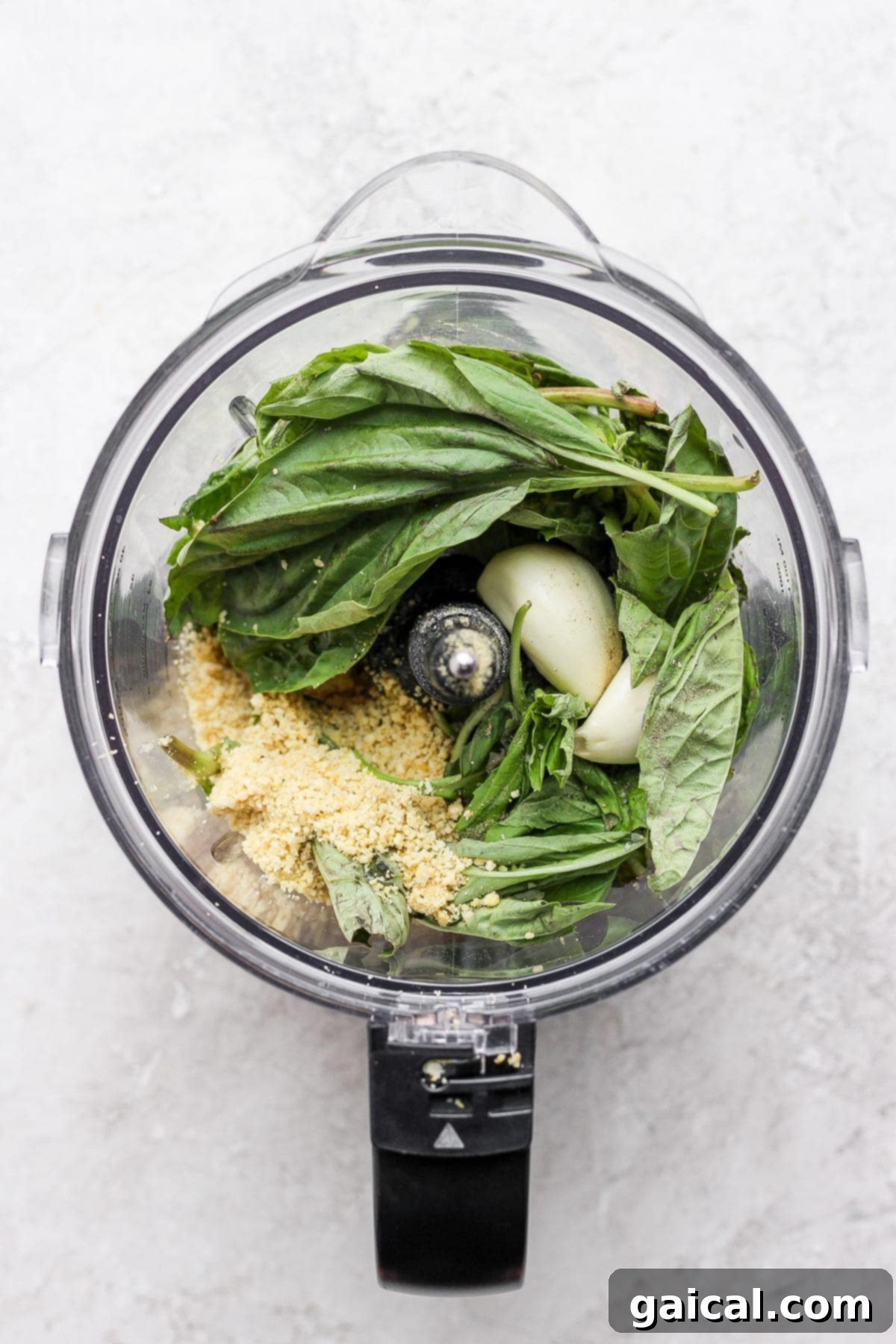 Fresh basil leaves, garlic, and other vegan pesto ingredients being added to a food processor.