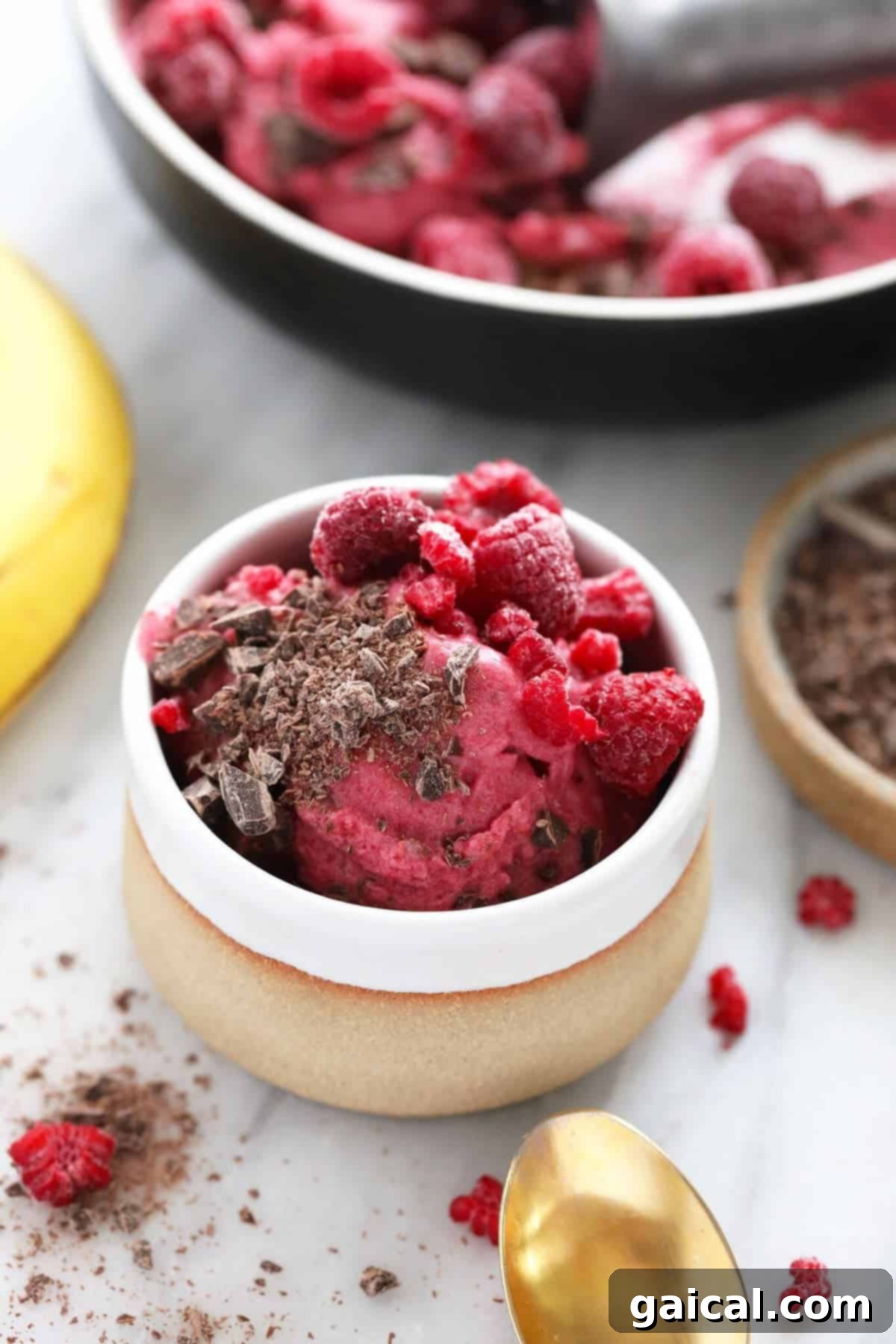 A bowl of raspberry nice cream topped with chocolate shavings and whole raspberries, with a banana and spoon nearby on a marble surface.