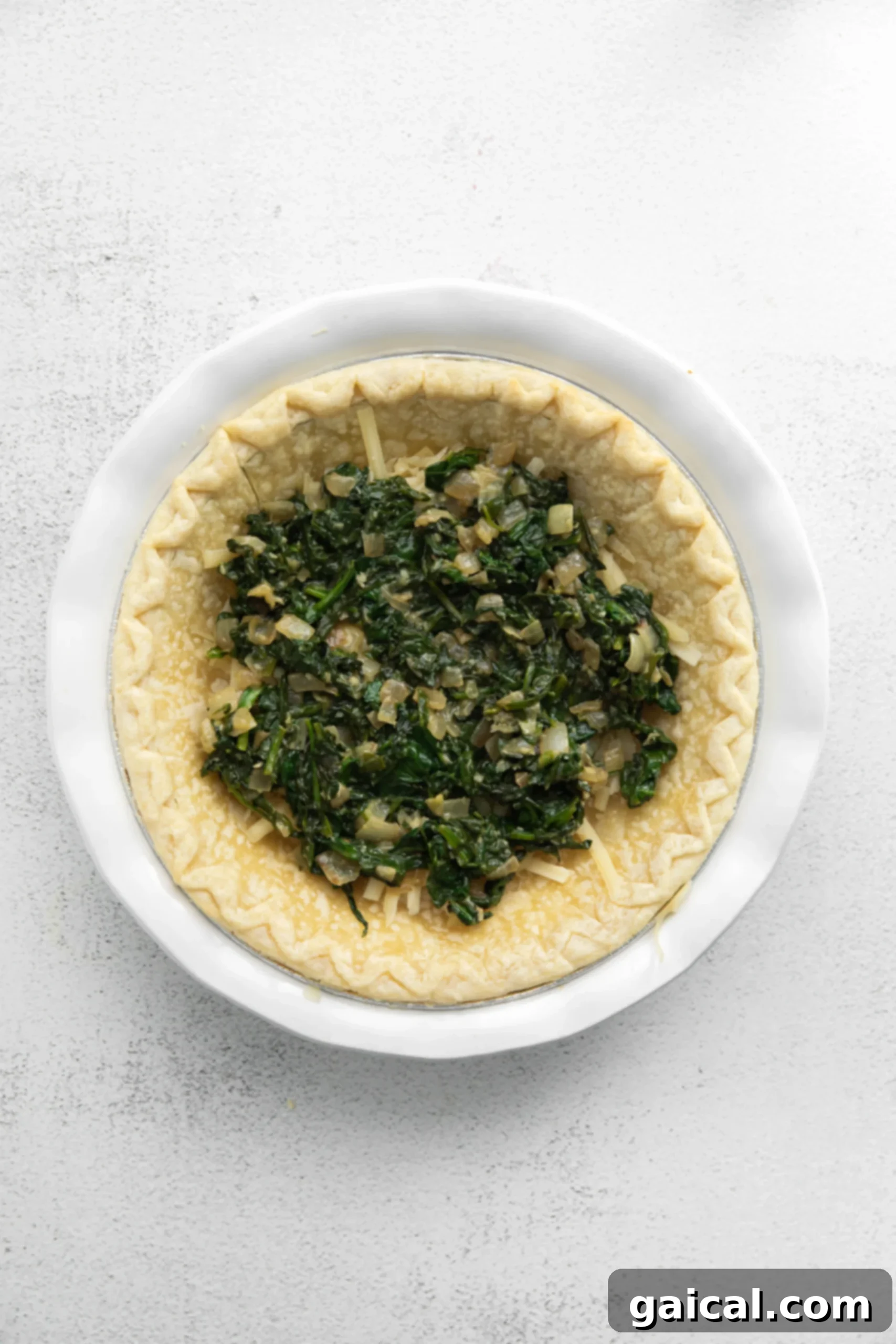 Simple & Savory Spinach Quiche 5 Fresh spinach, translucent minced white onions, and fragrant minced garlic cloves are artfully arranged in a par-baked pie crust, showcasing the vibrant and aromatic base of the quiche filling.