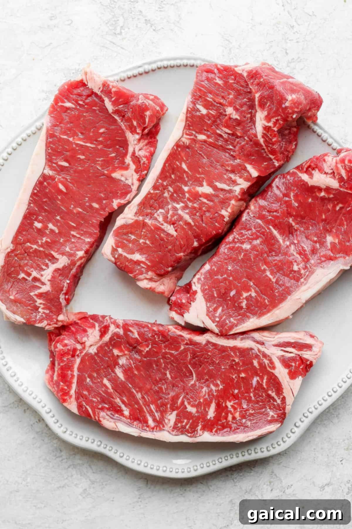 Four large, uncooked New York strip steaks on a plate, ready for seasoning and grilling.