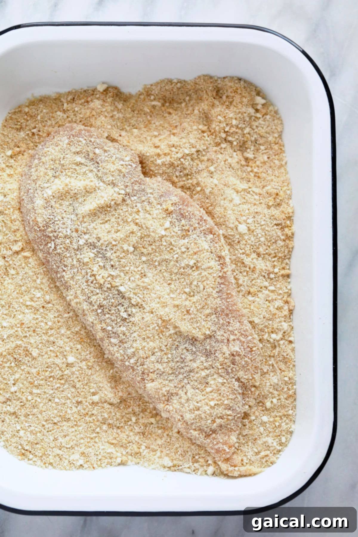 Chicken cutlet being dredged in a bowl filled with a mixture of bread crumbs and grated Parmesan cheese.