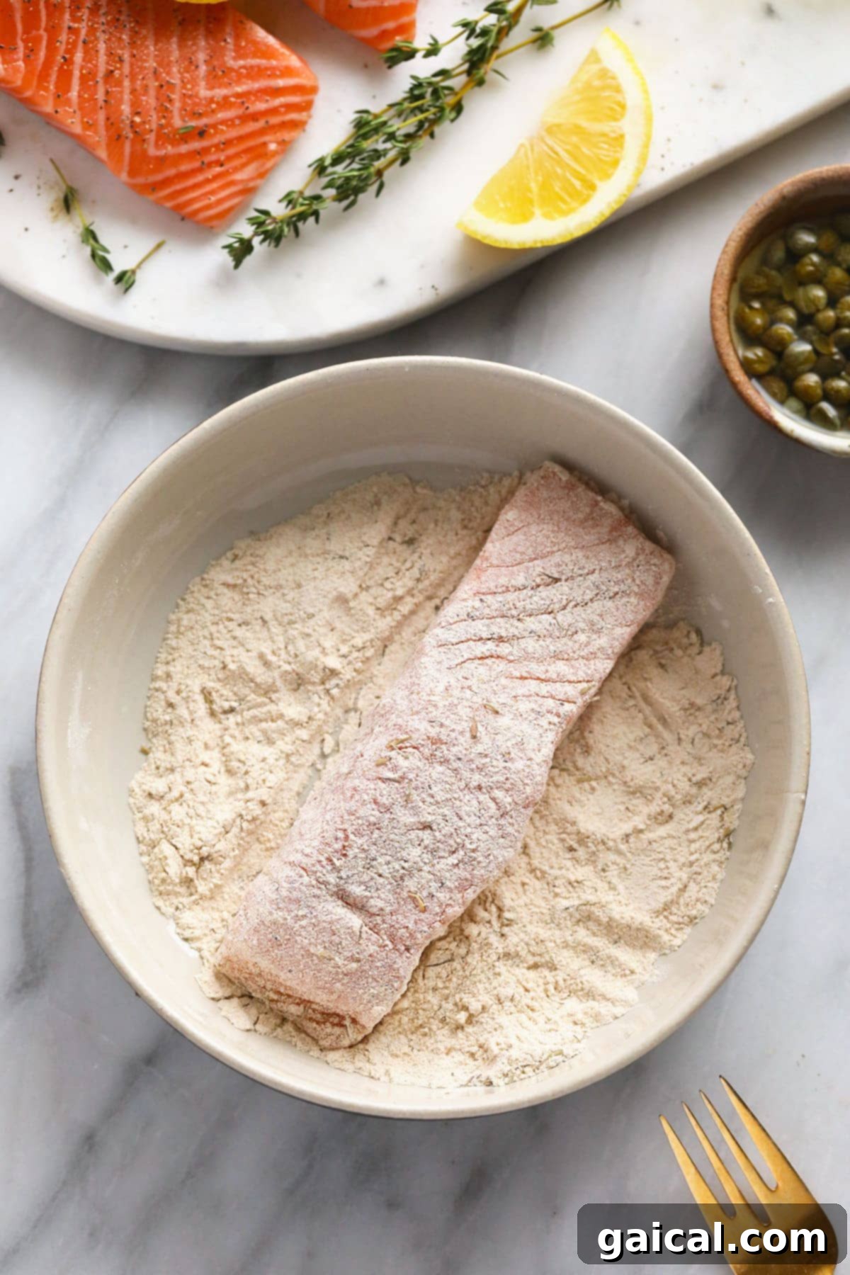 Salmon fillet being dredged through a mixture of flour and spices in a shallow bowl before searing.