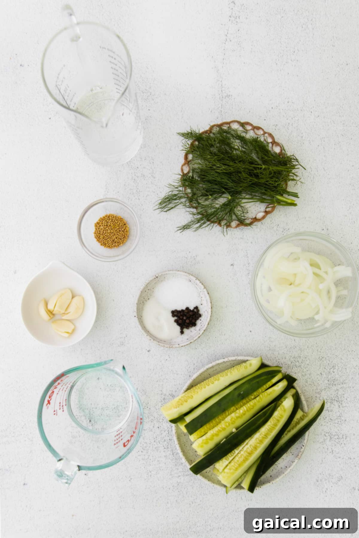 Sliced cucumbers, fresh dill sprigs, white onion slices, mustard seeds, sugar, garlic cloves, and white vinegar neatly arranged on a countertop, ready for pickling.