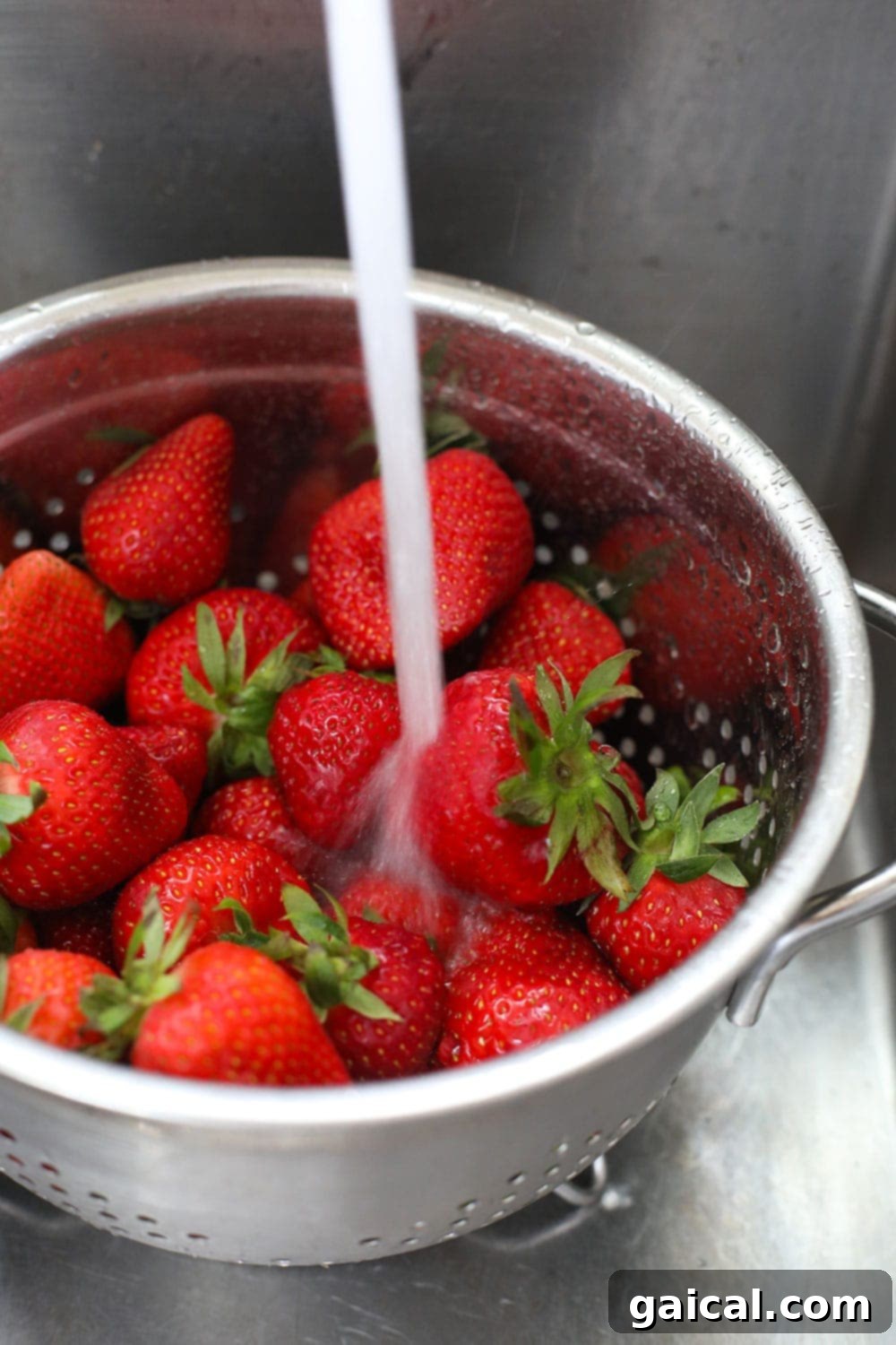 Fresh strawberries being rinsed under cold running water in a colander before preparation for freezing, showing clear water draining.