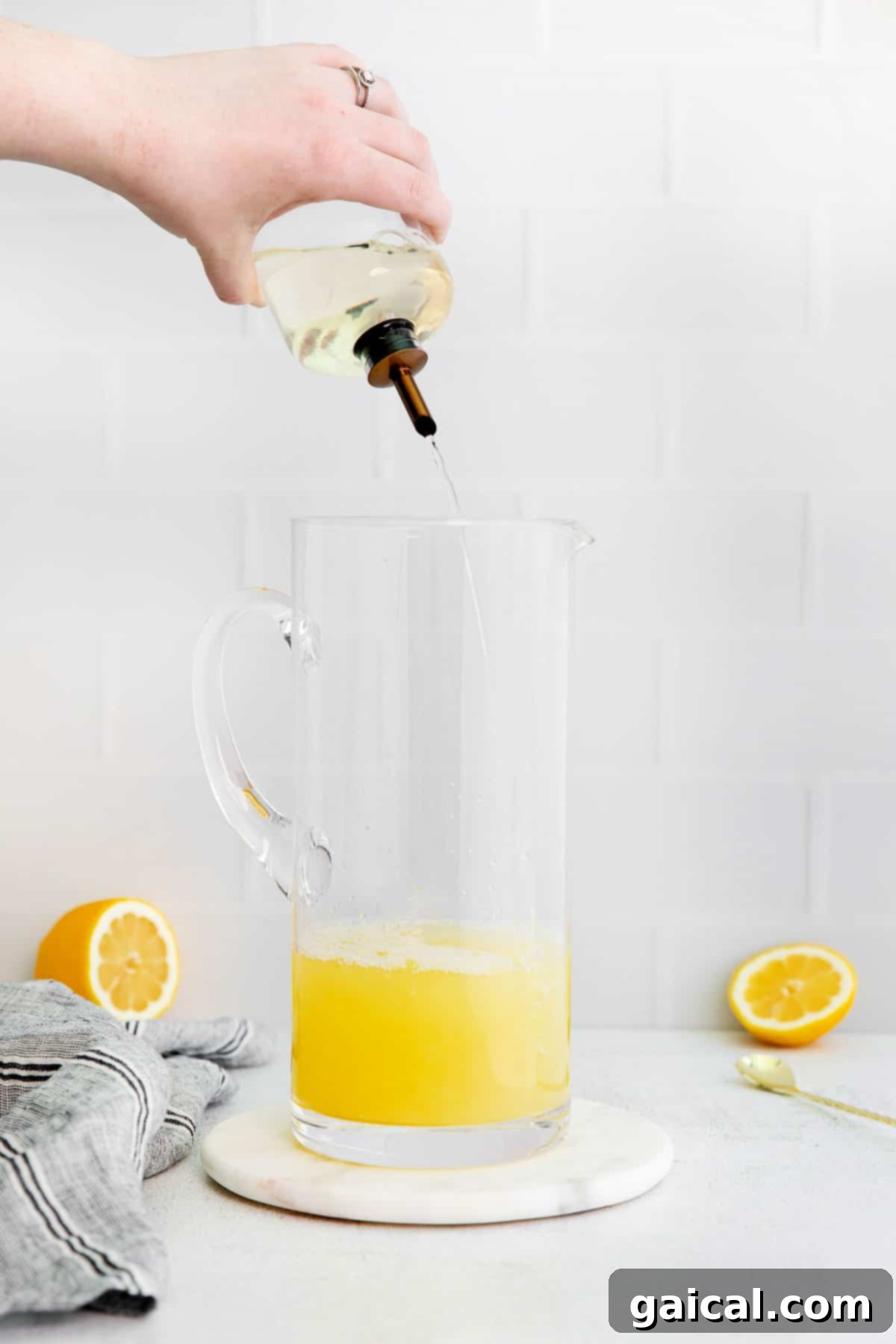 A stream of golden simple syrup being poured into a pitcher of fresh lemon juice and water, illustrating the blending process.