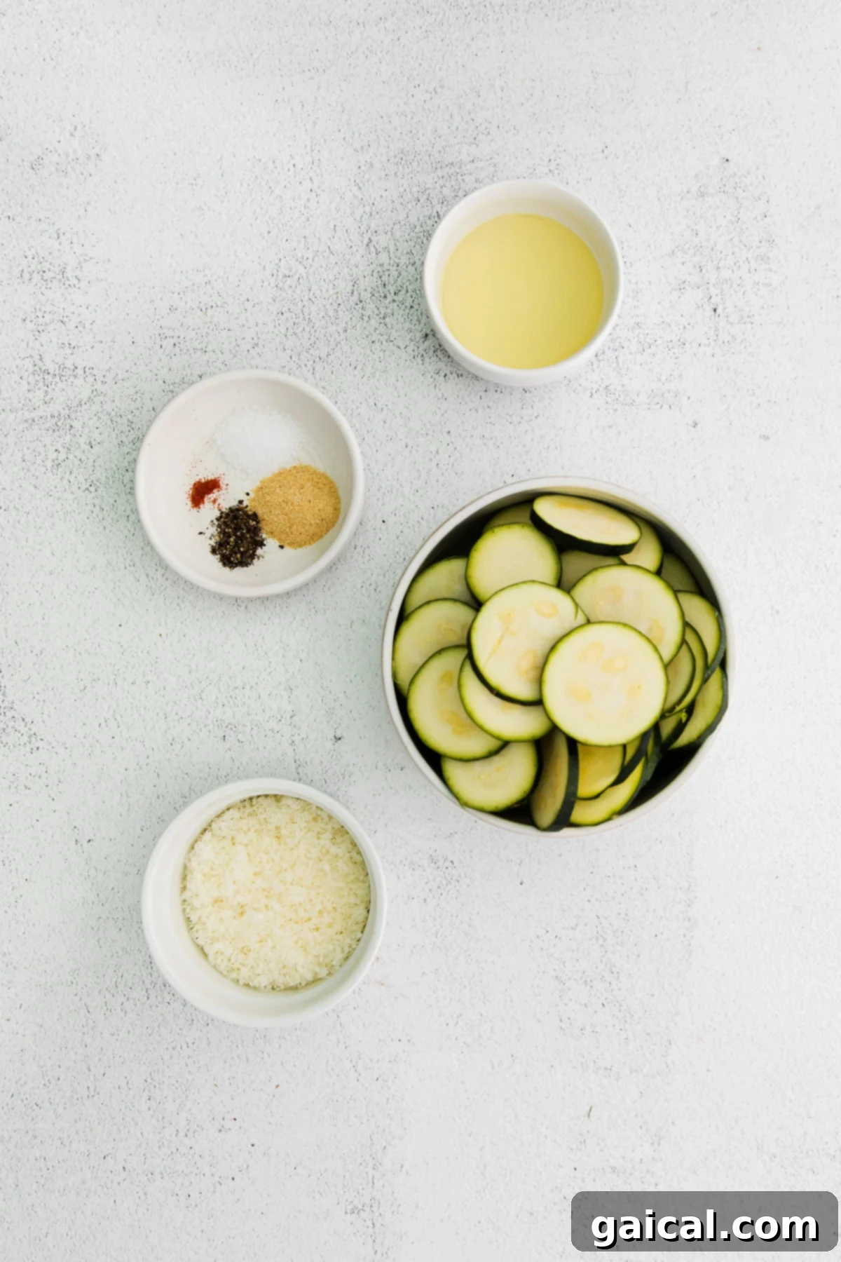 Crispy Parmesan Zucchini Bites 3 A beautifully arranged collection of fresh ingredients for baked zucchini in small, rustic bowls: olive oil, precisely sliced zucchini rounds, a pile of finely grated Parmesan cheese, a small bowl of garlic powder, freshly ground black pepper, a tiny pinch of cayenne pepper, and sparkling sea salt.