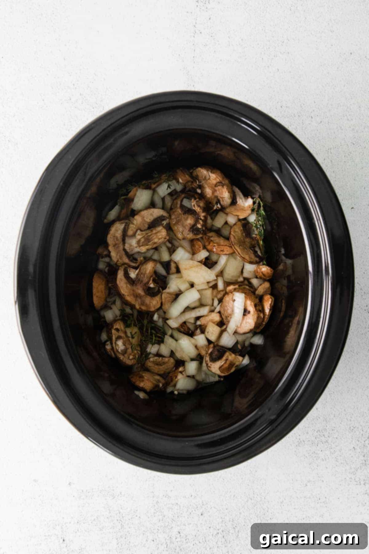 A colorful array of sliced mushrooms, diced onions, and fresh thyme sprigs resting in the bottom of a slow cooker, ready for the beef.