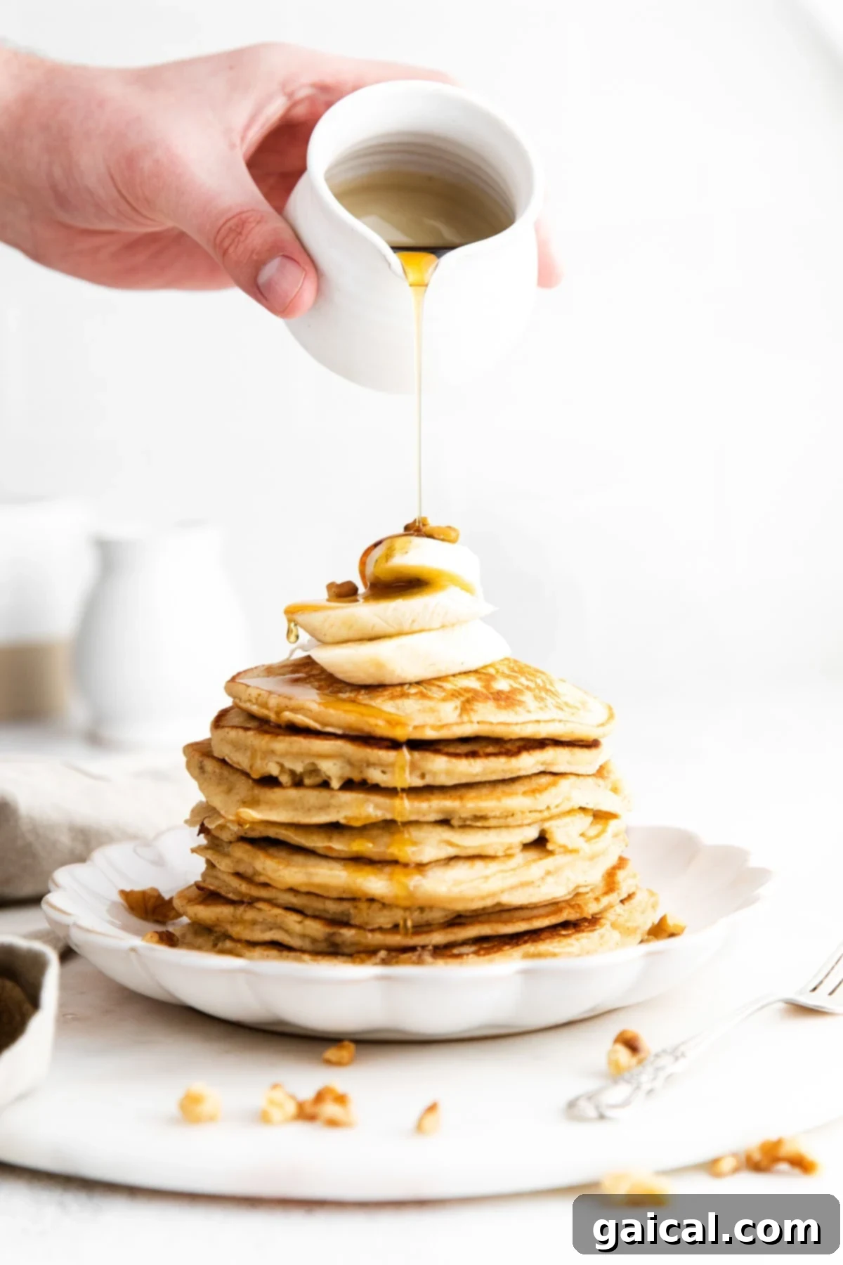 Nourishing Banana Pancakes 2 Golden brown banana pancakes being drizzled with warm maple syrup, garnished with fresh banana slices.