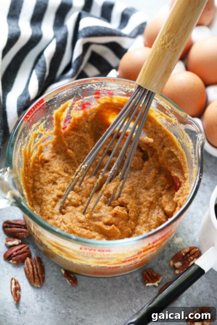 A close-up shot of vibrant orange pumpkin pancake batter being vigorously whisked in a clear glass bowl, showcasing its smooth texture and readiness for the skillet.