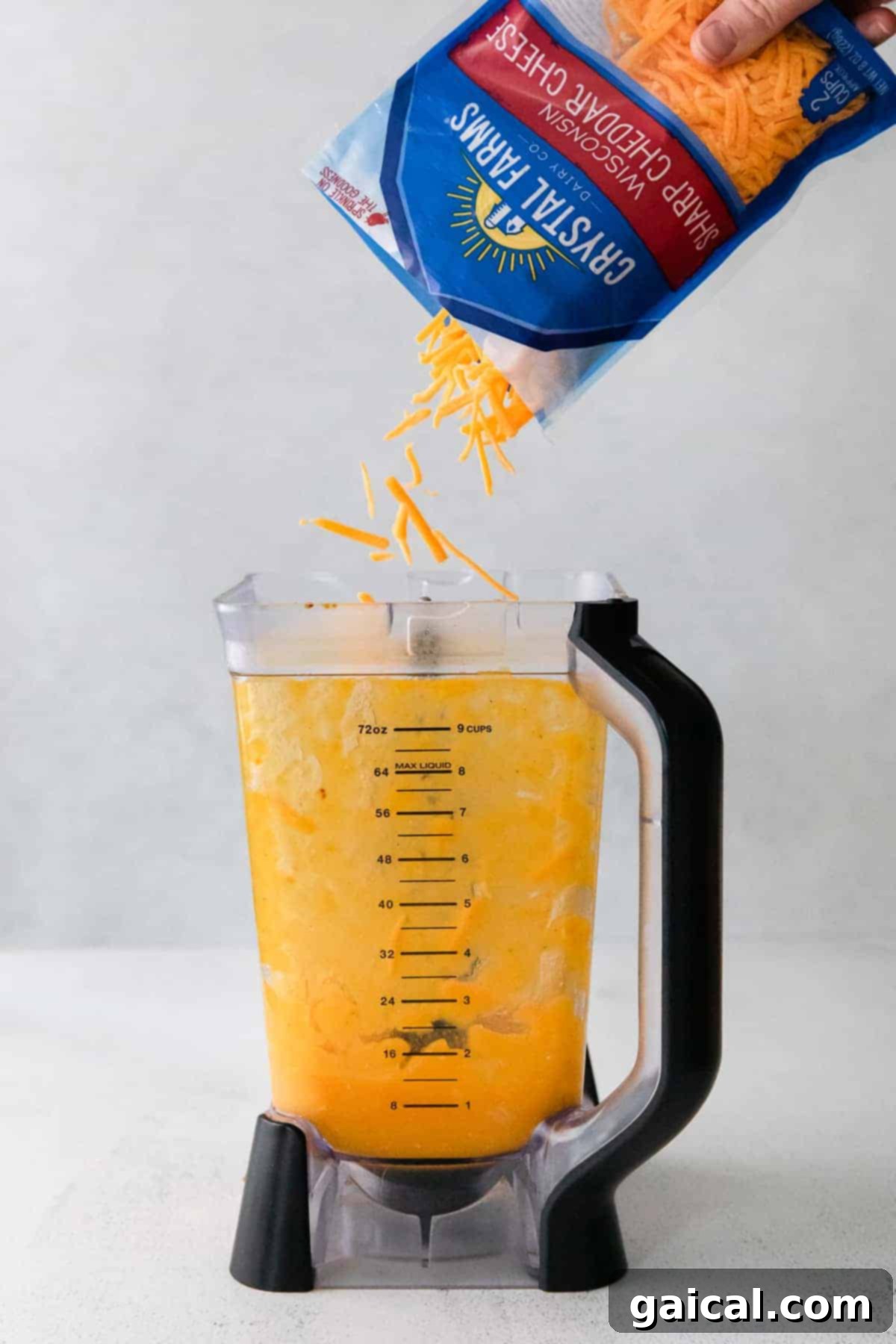 Golden Butternut Mac 4 Shredded cheddar cheese being carefully poured into a blender with roasted vegetables.
