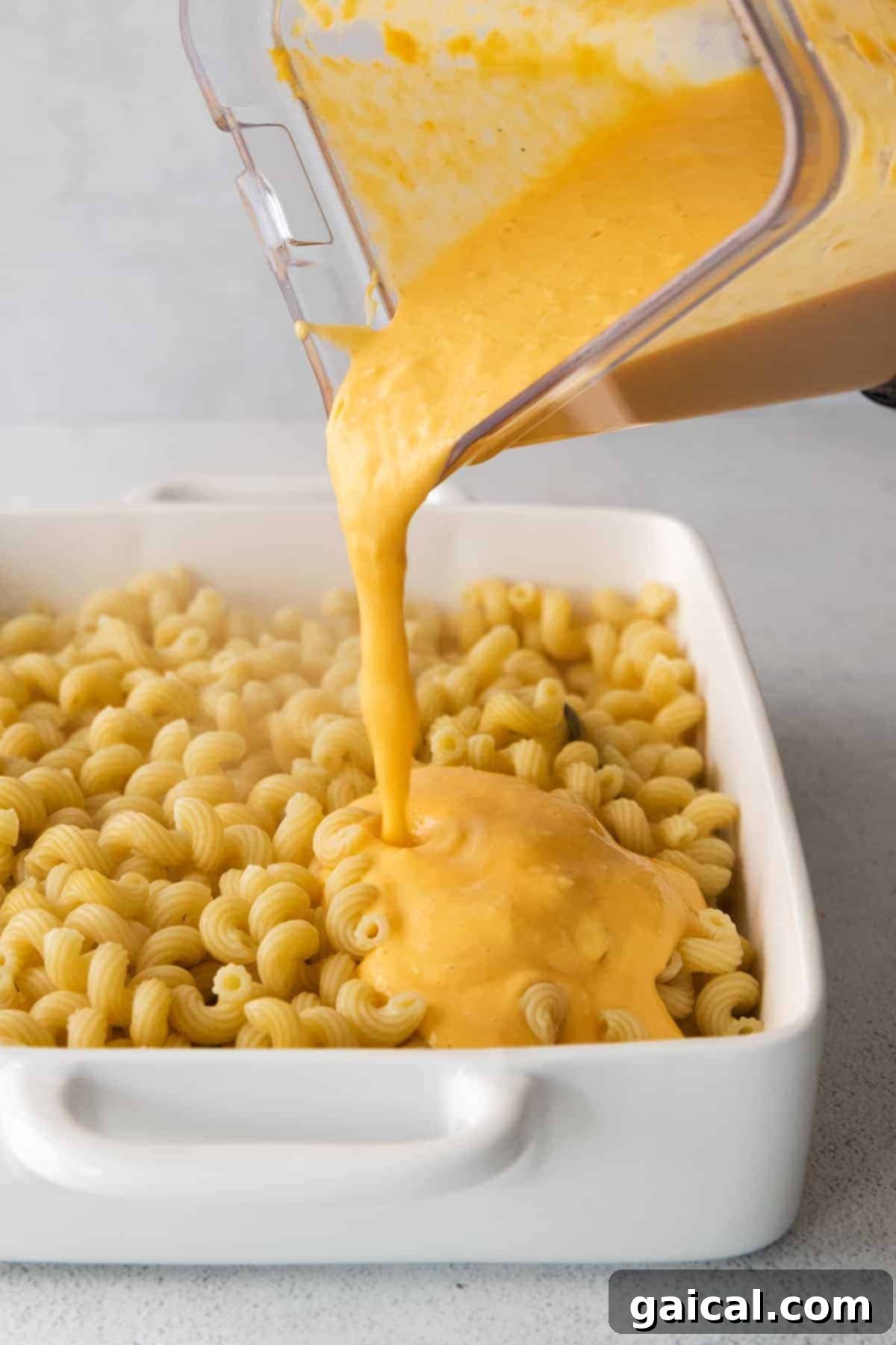 Golden Butternut Mac 5 Smooth butternut squash cheese sauce being generously poured over cooked cavatappi noodles in a baking dish.
