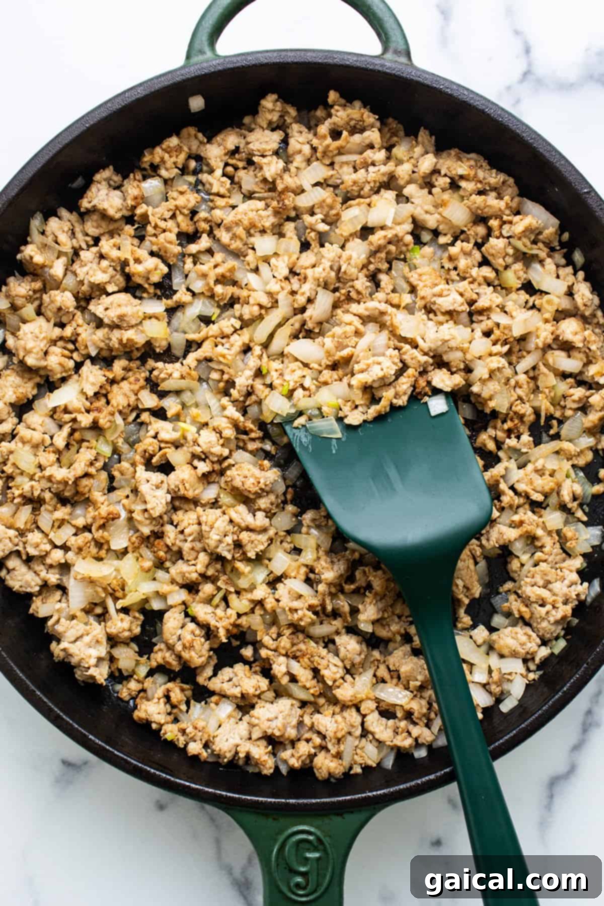 Vibrant Healthy Enchilada Dip 3 Ground chicken cooked with diced onions in a cast iron skillet, being stirred with a green spatula.