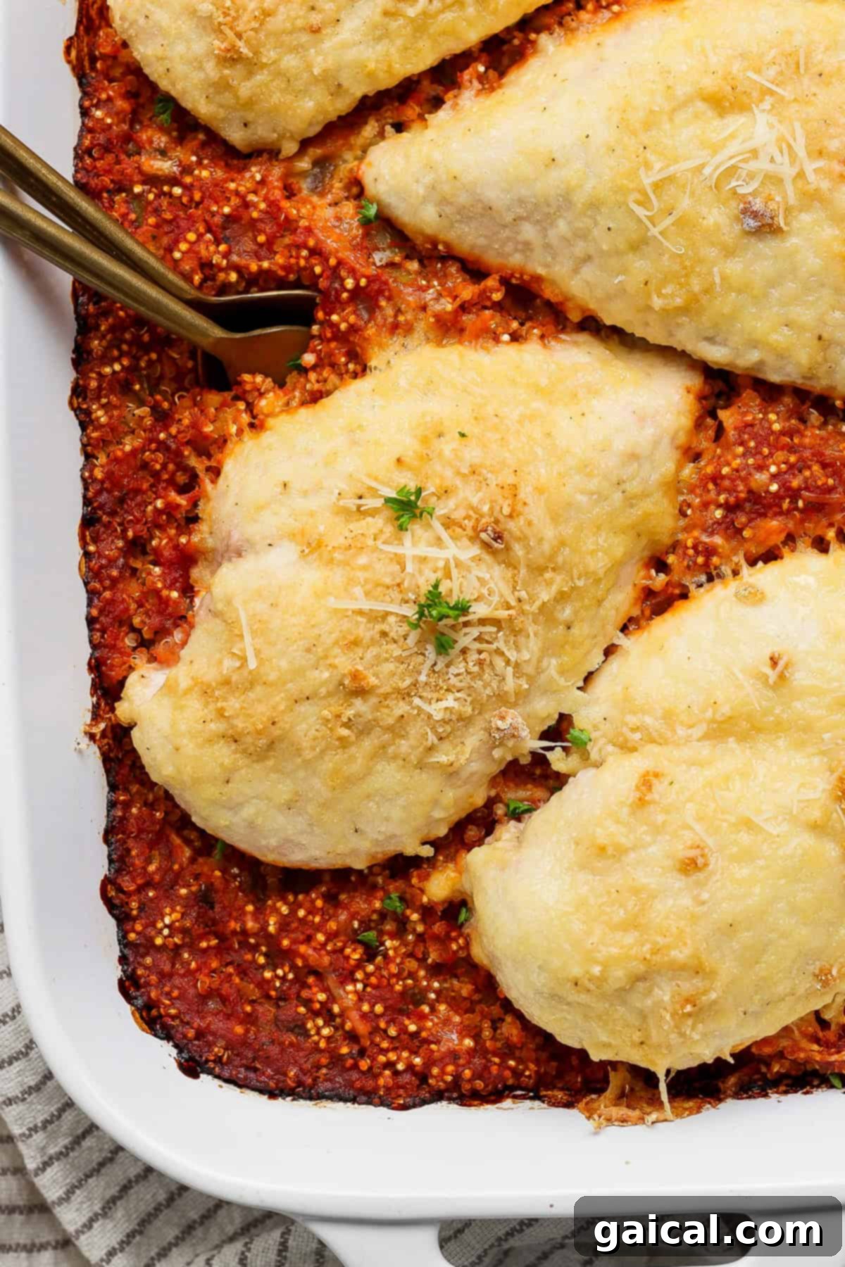 Parmesan Chicken Quinoa Bake 2 Baked chicken breasts topped with perfectly melted golden cheese on a bed of vibrant red tomato quinoa in a pristine white baking dish, elegantly garnished with fresh green herbs and additional grated cheese.