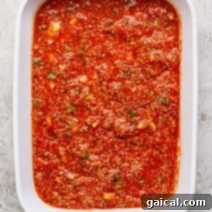 A close-up view of a white baking dish filled with a vibrant red tomato sauce base, rich with visible herbs and spices, mixed with quinoa, creating a hearty foundation for the casserole.