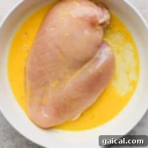 A raw chicken breast immersed in a white bowl containing beaten eggs, resting on a gray textured kitchen surface, illustrating the essential egg wash step before breading.