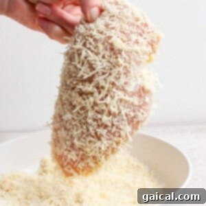 A hand gently patting grated cheese and flour onto a raw chicken breast, ensuring a thorough and even coating over a white bowl filled with the breading mixture, ready for baking.