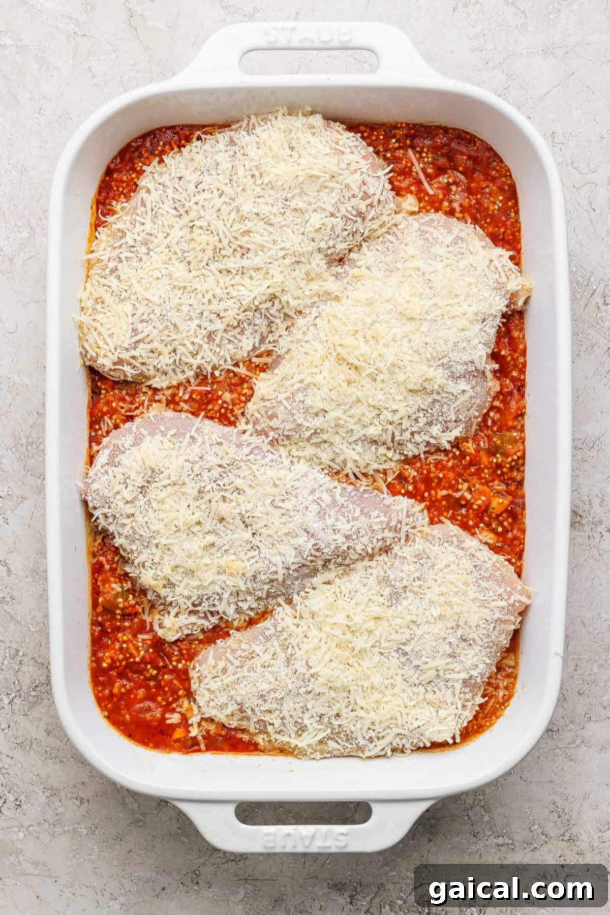 Parmesan Chicken Quinoa Bake 5 Four beautifully breaded, uncooked chicken breasts, generously covered in grated parmesan cheese, are meticulously placed on top of a bubbling layer of rich tomato sauce and quinoa in a white oven-safe baking dish, prepared for the next stage of baking.
