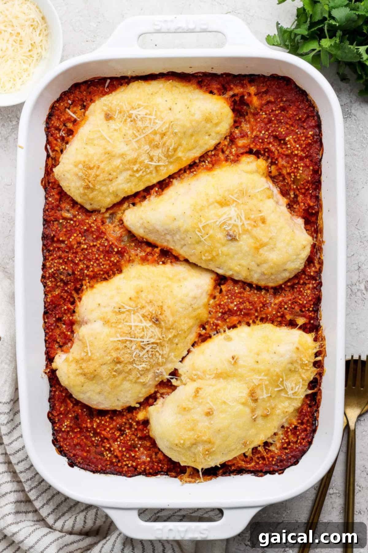 Parmesan Chicken Quinoa Bake 6 A beautifully baked chicken parmesan quinoa casserole in a white baking dish, featuring golden-brown, cheesy chicken breasts resting on a rich red sauce base, presented with a serving fork and a neatly folded linen napkin beside it, ready for a comforting meal.