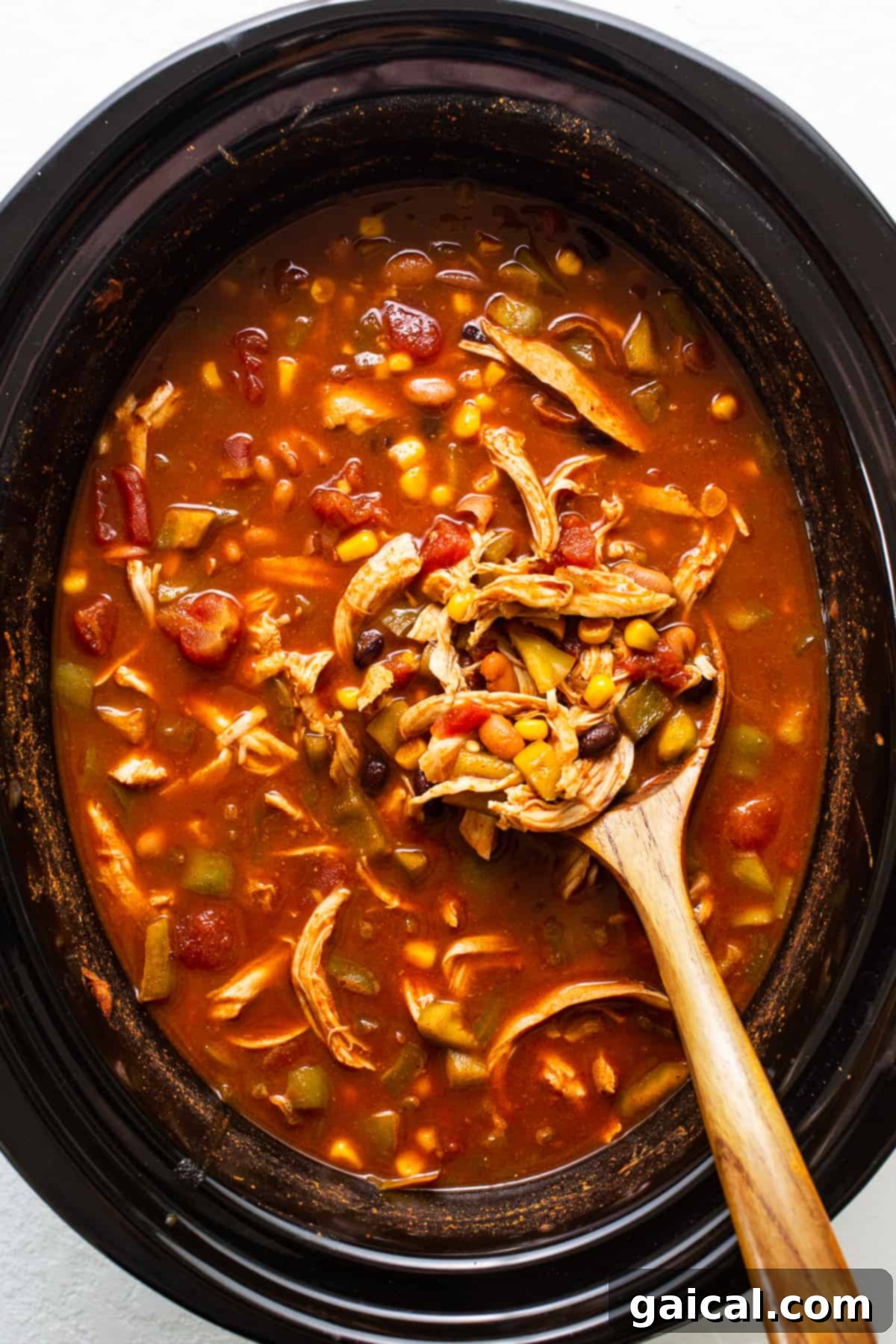 A wooden ladle scooping a generous portion of chicken enchilada soup from the slow cooker, showing shredded chicken and vegetables.