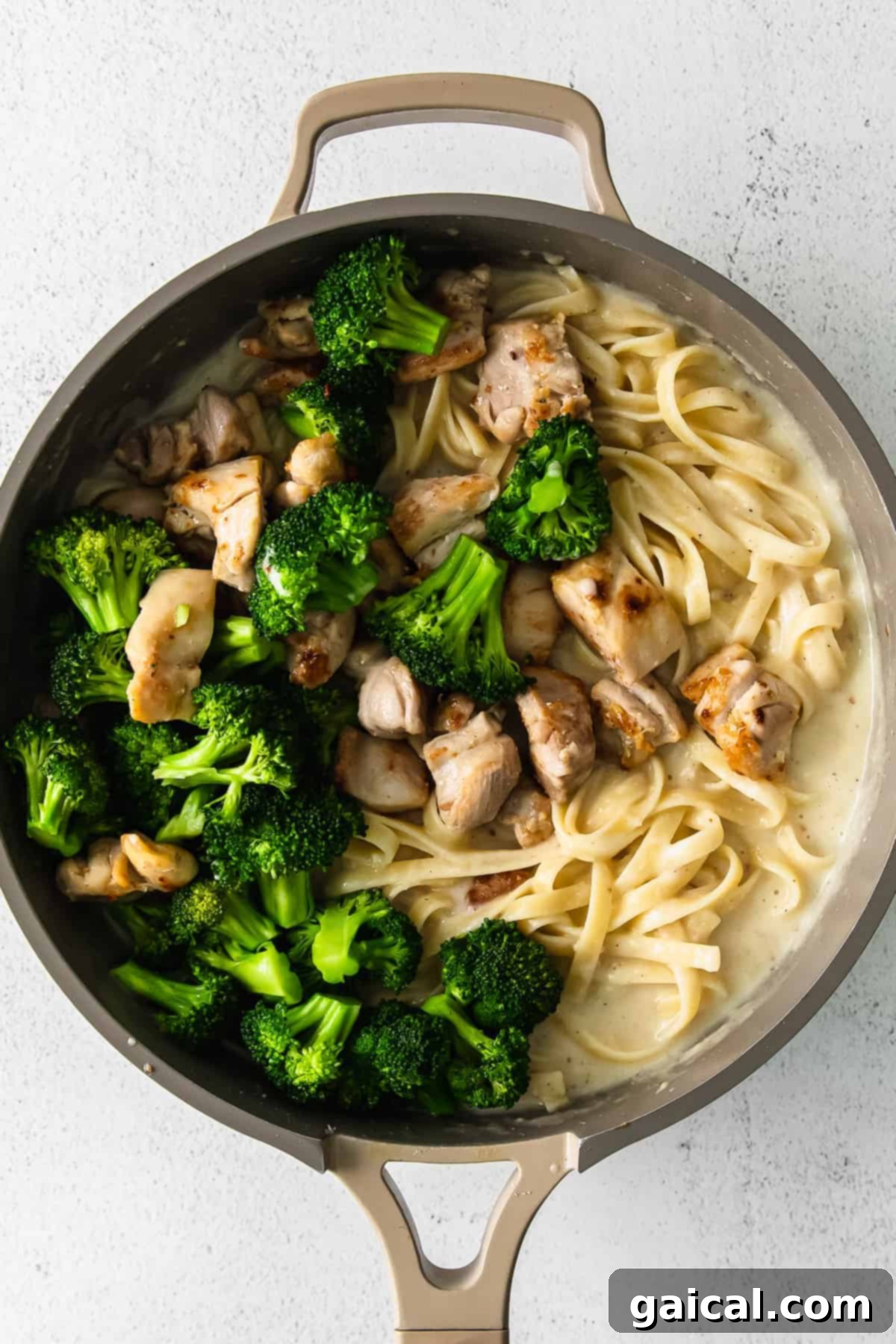 Cooked fettuccine noodles, steamed broccoli, and browned chicken thighs mixed in a skillet with alfredo sauce.