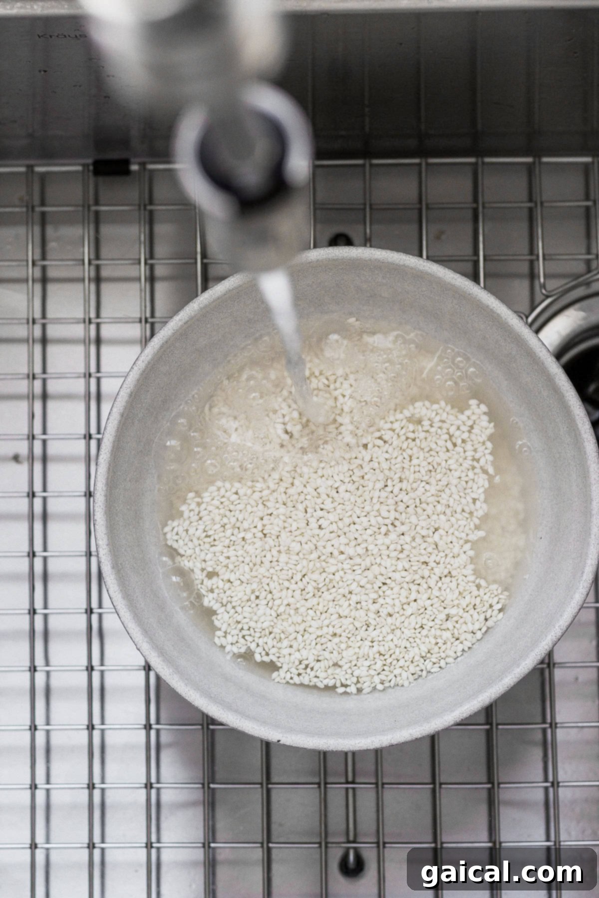 Hands rinsing glutinous sweet rice in a bowl under warm running water in a kitchen sink.