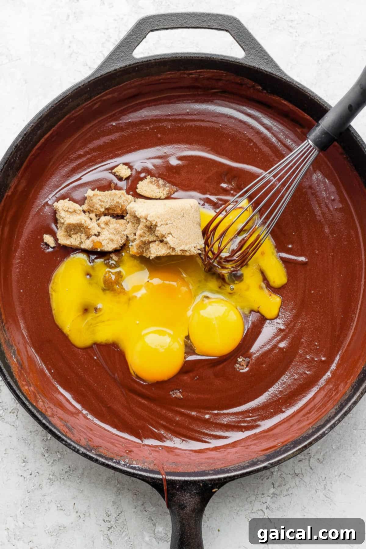 Combining Ingredients in Skillet Egg yolks, light brown sugar, and semi-sweet chocolate chips being added to the melted chocolate and butter mixture in a skillet.