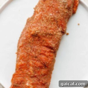 A raw pork tenderloin generously coated with a savory dry rub, placed on a white cutting board.