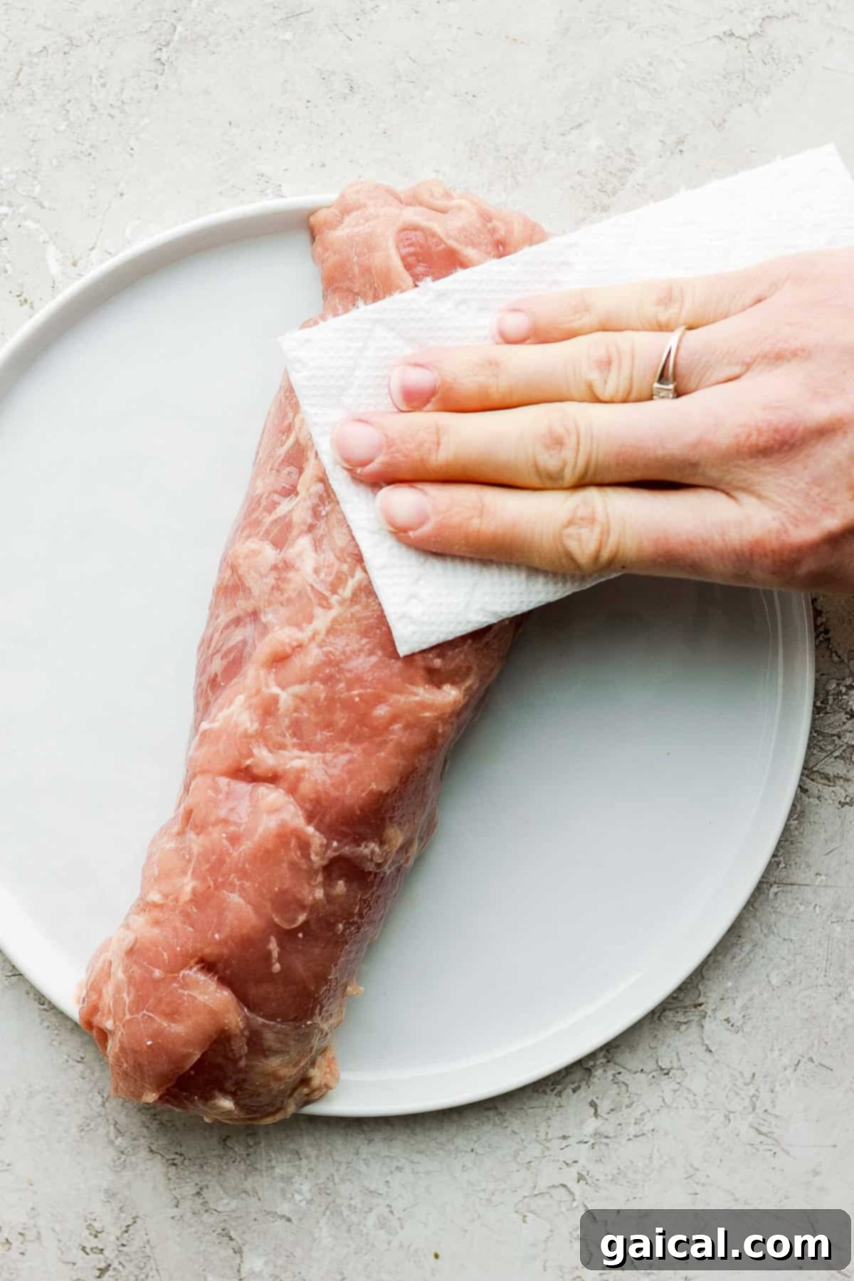 This Air Fryer Pork Tenderloin recipe is super moist on the inside with a crispy outside and is ready to enjoy in minutes. A person is patting a raw pork tenderloin dry with a paper towel on a white plate, preparing it for seasoning.