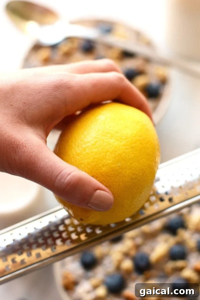 Adding Fresh Lemon Zest to Blueberry Overnight Oats Close-up of hands zesting a fresh lemon over a bowl, highlighting the vibrant yellow peel and the aromatic process of adding citrus flavor.
