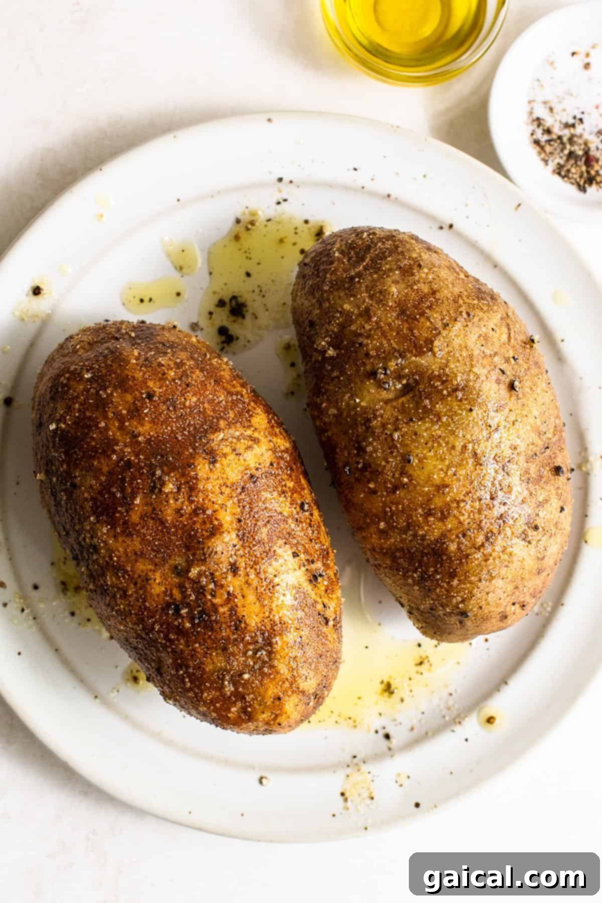 Raw russet potatoes, olive oil, and a small bowl of salt and pepper laid out on a kitchen surface, ready for preparation.