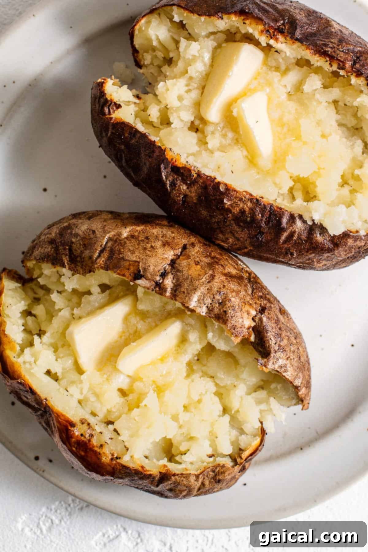 A split air fryer baked potato topped with a generous pat of melting butter and a sprinkle of fresh herbs.