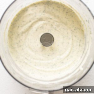A food processor bowl containing a smooth, vibrant green mixture, indicating the initial blending of the creamy ingredients and fresh dill.