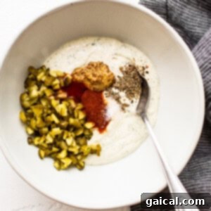 A bowl of creamy dip infused with green flecks of dill and minced pickles, being stirred with a spoon, highlighting the texture before salmon is added.
