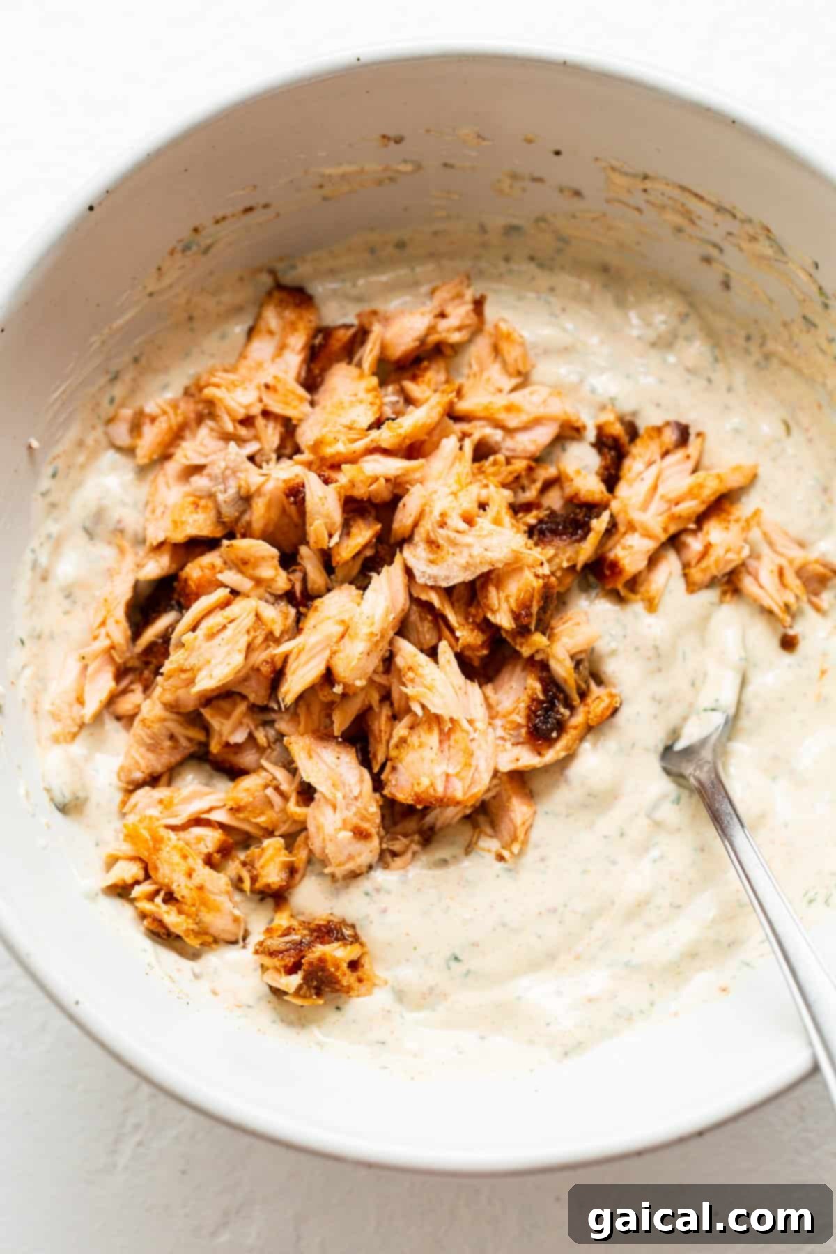 Incorporating Flaked Smoked Salmon for Optimal Texture A close-up of a white bowl with a creamy mixture, now infused with generous flakes of pink smoked salmon, being gently mixed with a spoon, showing the final stages of preparation.