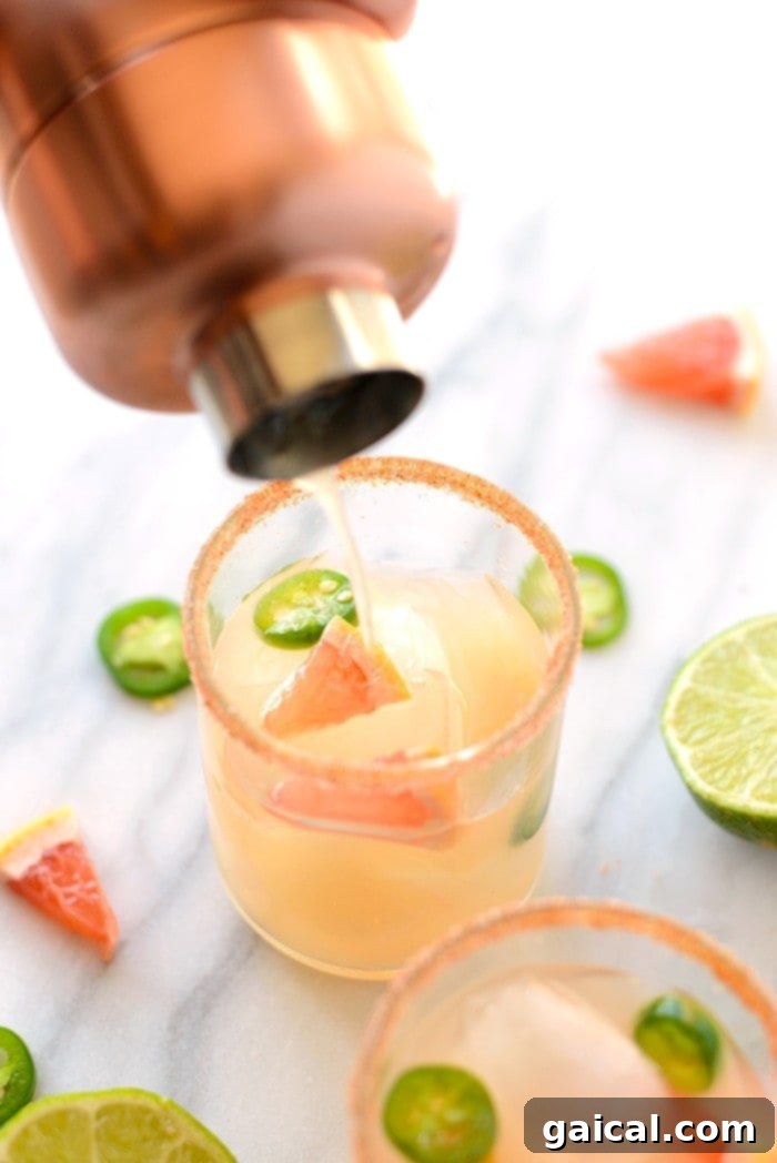 Chili-Kissed Grapefruit Margarita 3 A hand pouring a grapefruit margarita from a cocktail shaker into a salt-rimmed glass filled with ice.